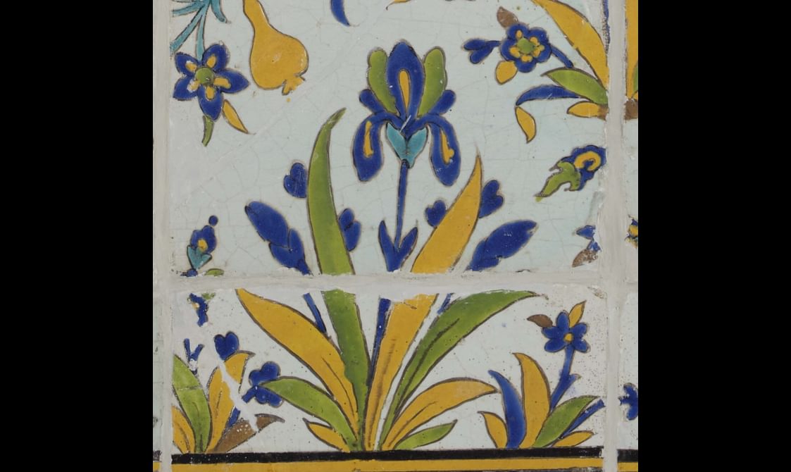 A section of a long, rectangular panel made of hundreds of intricately decorated, painted tiles. This section focuses on a small plant with flowers. The colours painted are yellow, blue, green and brown.