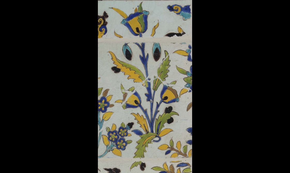 A section of a long, rectangular panel made of hundreds of intricately decorated, painted tiles. This section focuses on a small plant with flowers. The colours painted are yellow, blue, green and brown.
