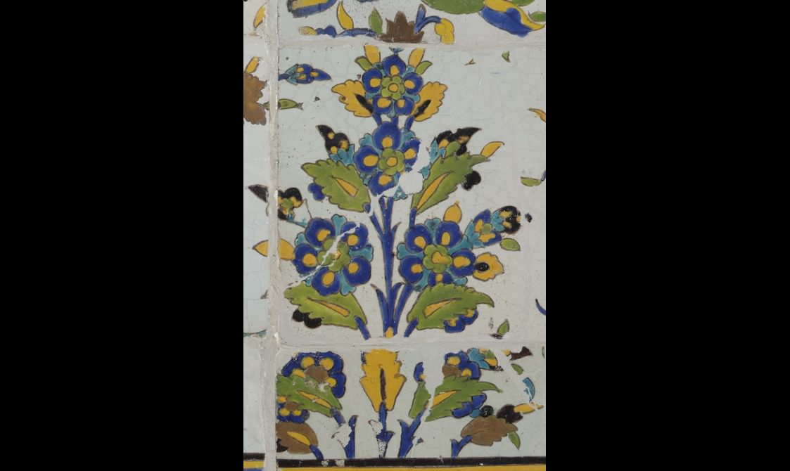 A section of a long, rectangular panel made of hundreds of intricately decorated, painted tiles. This section focuses on a small plant with flowers. The colours painted are yellow, blue, green and brown.