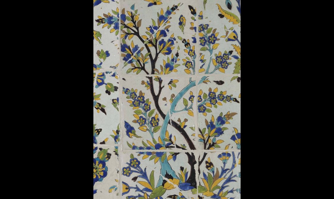 A section of a long, rectangular panel made of hundreds of intricately decorated, painted tiles. This section focuses on a tree. The colours painted are yellow, blue, green and brown.