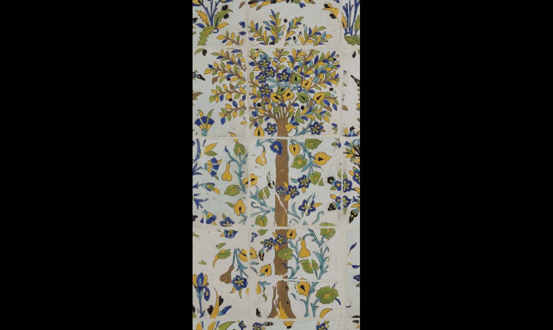 A section of a long, rectangular panel made of hundreds of intricately decorated, painted tiles. This section focuses on a tall tree. The colours painted are yellow, blue, green and brown.
