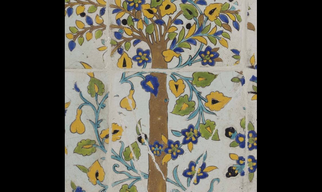 A section of a long, rectangular panel made of hundreds of intricately decorated, painted tiles. This section focuses on a small plant with flowers. The colours painted are yellow, blue, green and brown.