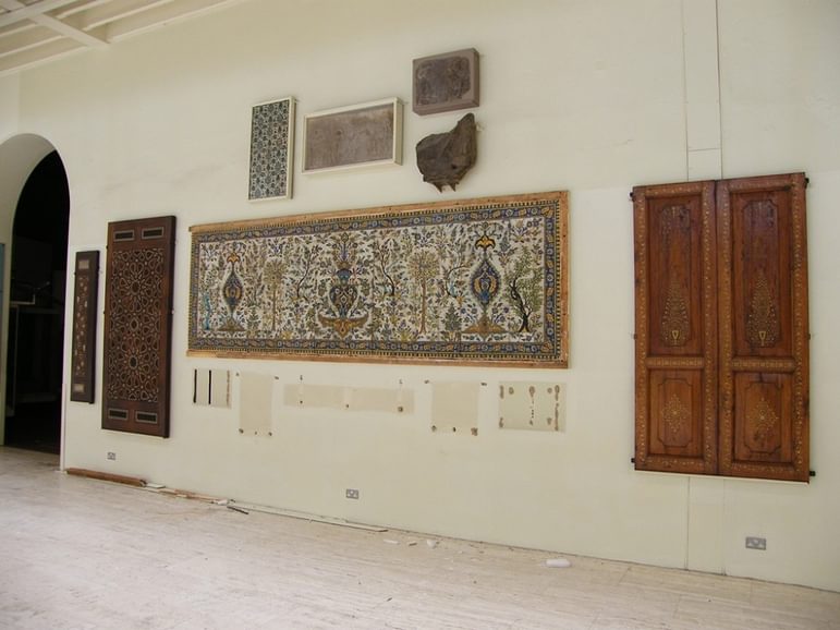 The Iranian tile panel, which is long and rectangular, and made up of painted tiles in blue, green, brown and yellow, is mounted on a wall in a museum gallery.