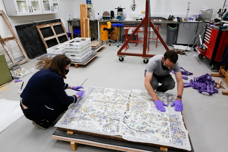 Two conservatiors are wearing masks and purple gloves, and are in a workshop. They are kneeling on the ground, painting and restoring the Iranian tile panel which is slightly raised.