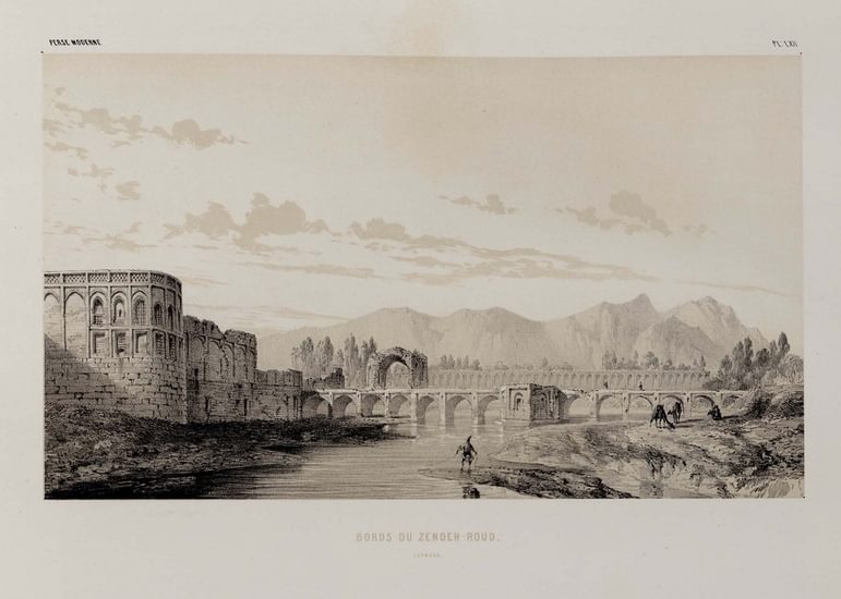 A black and white image of the outside of a palace. The edge of the palace is in view and rounded, and you can see a bridge with arched supports going over a body of water surrounding the palace. A person stands on the bank, with what looks like a camel behind. In the distance are mountains.