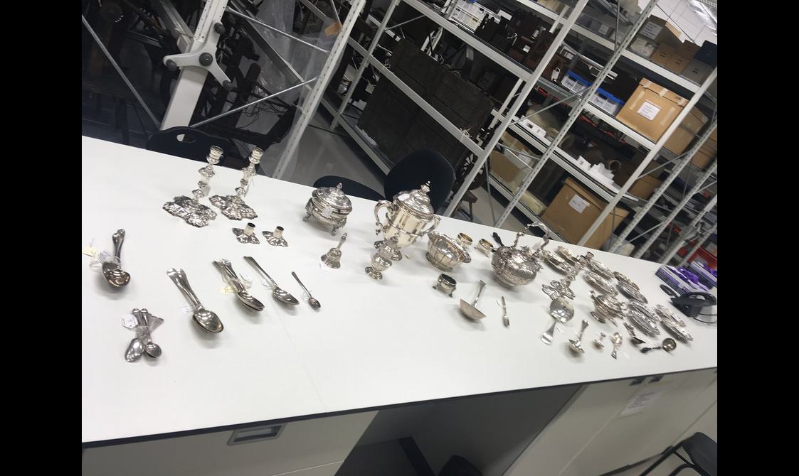 A long white table with several silver objects on top including spoons, candles sticks, and teapots. There are industrial looking shelves in the background stacked with boxes and objects.
