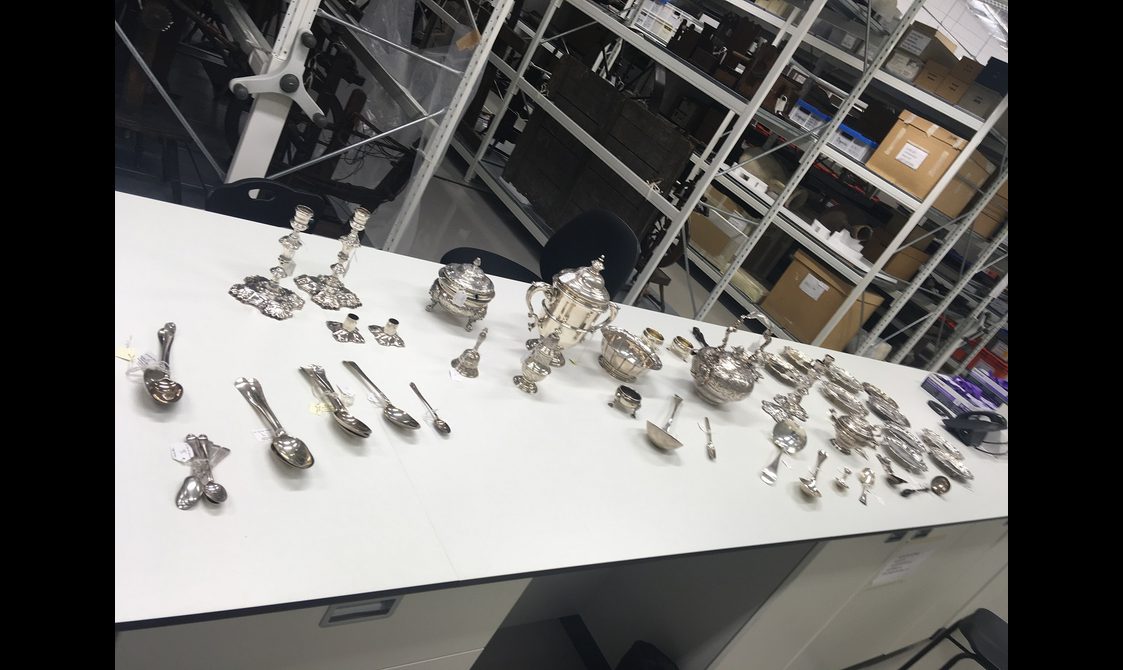 A long white table with several silver objects on top including spoons, candles sticks, and teapots. There are industrial looking shelves in the background stacked with boxes and objects.