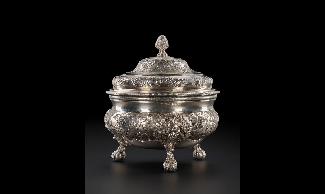 An ornate silver sugar bowl with three short legs and a lid