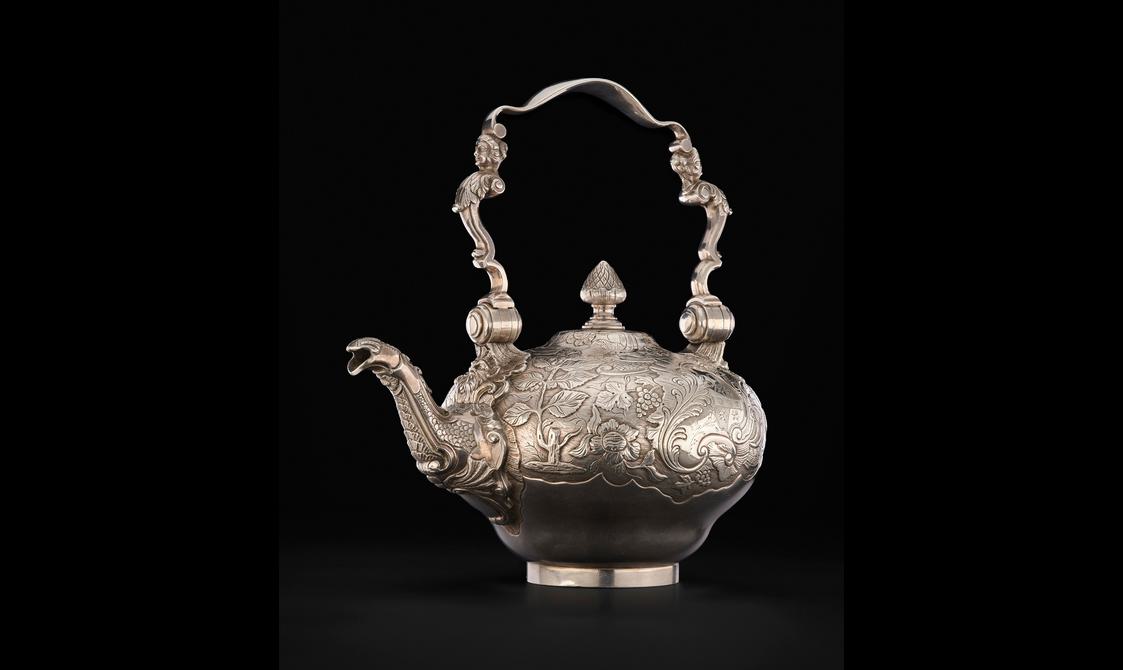 A silver tea kettle decorated with detailed floral designs. It has a tall handle which depicts a human face on each side.