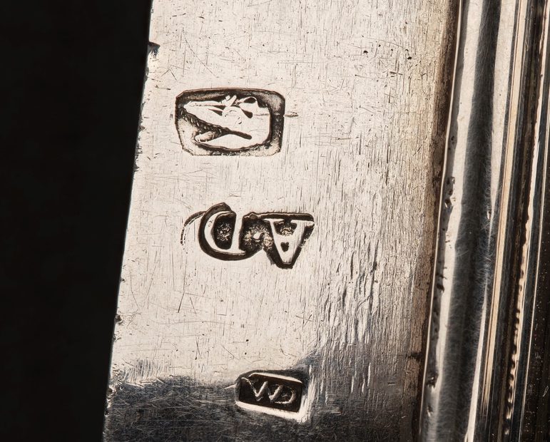 A detail of a silver object showing stamps on its surface. One stamp has the initials A D and the other marks are more obscure.