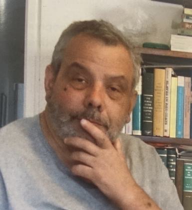 A man with short hair and a beard wearing a grey tshirt with one hand on his chin. There are bookshelves in the background.