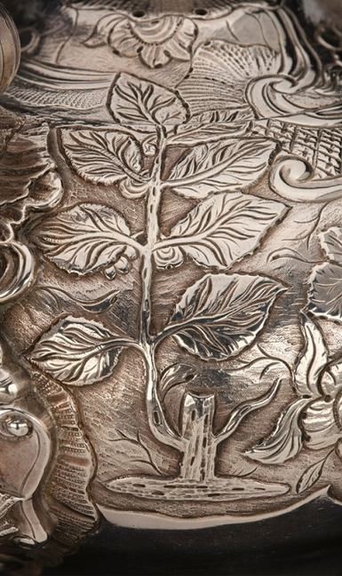 A detail of a silver object with a tree and leaves on the surface