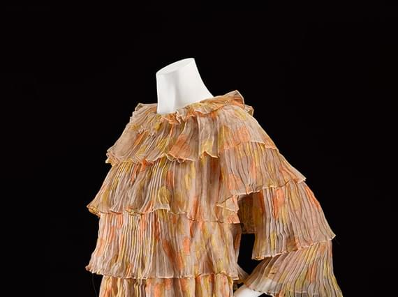 A mannequin wearing a beige and orange patterned mini dress with tiered silk chiffon layers and three quarter length sleeves.
