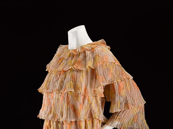 A mannequin wearing a beige and orange patterned mini dress with tiered silk chiffon layers and three quarter length sleeves.