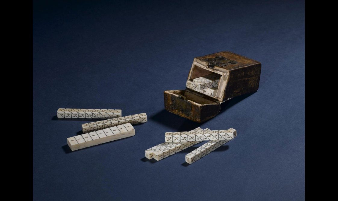 A colour photograph of some ivory sticks with numbers enranved on them and a leather case, against a dark blue background.