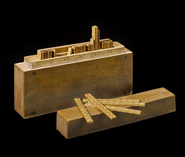 A colour photograph of a wooden box and lid containing some wooden sticks against a black background. The sticks have number pairs separated by diagonal lines.
