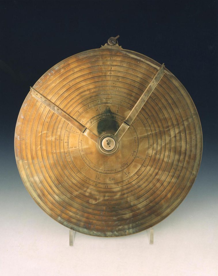 A colour photograph of a yellow metal circular device against a gradient background with two dials attached from the centre to the circumference.