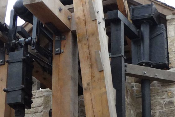 A detail of a large engine made of iron parts and wooden beams attached to a brick building.