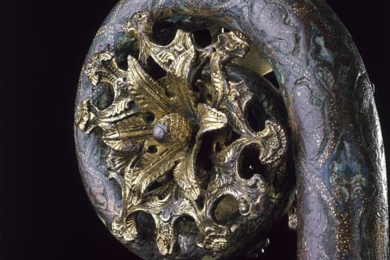 Closeup of metalwork object with a staff and a coiled top. The staff is grey-blue, and a golden sun-like burst is within its coiled top.