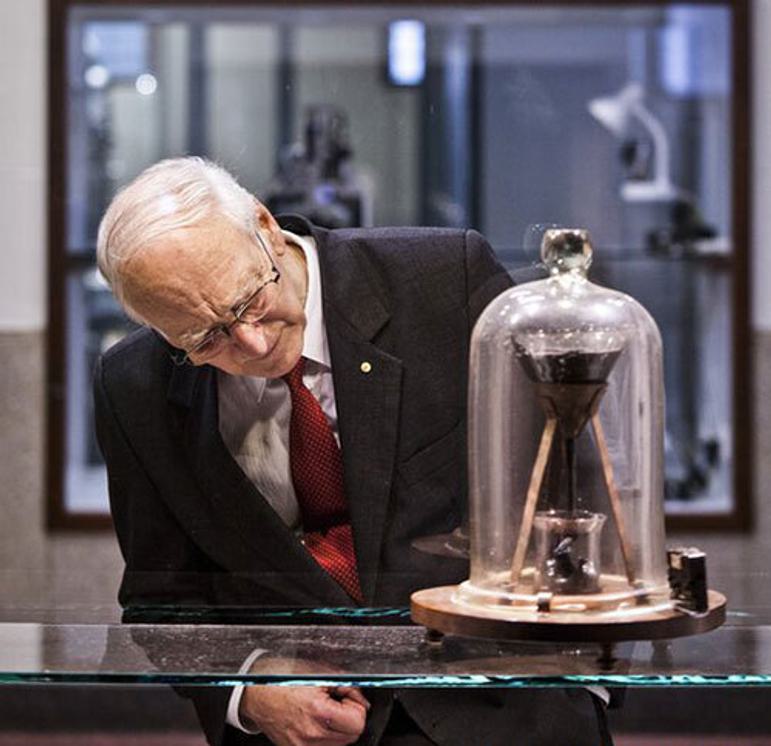 A man with white hair and glasses wearing a suit and tie looking into a bell jar that contains a funnels with a dark substance over a beaker.