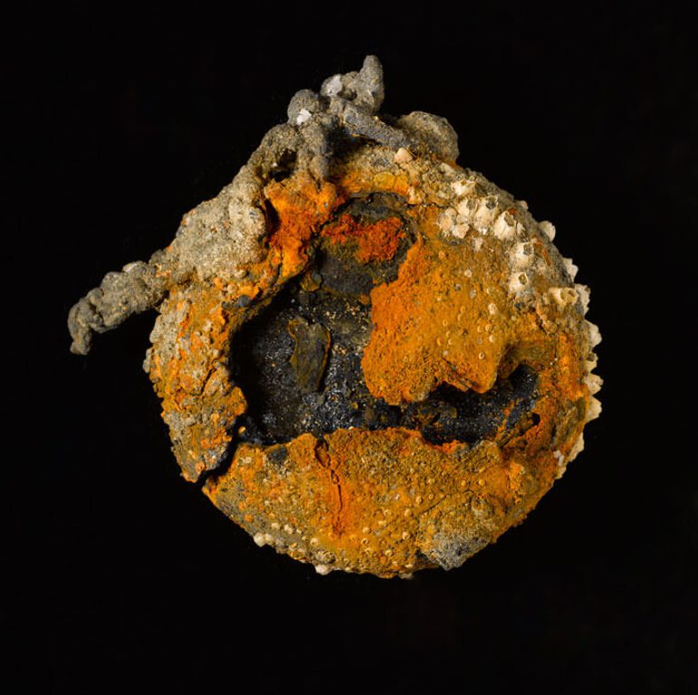 A colour photograph of a corroded circular piece of metal against a black background. The metal object has orange, black, and cream colouring, with the black at the centre. It is very bumpy in texture with what look like miniature barnacles attached to the outer circumference.