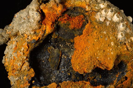 A colour photograph of a corroded circular piece of metal against a black background. The metal object has orange, black, and cream colouring, with the black at the centre. It is very bumpy in texture with what look like miniature barnacles attached to the outer circumference.
