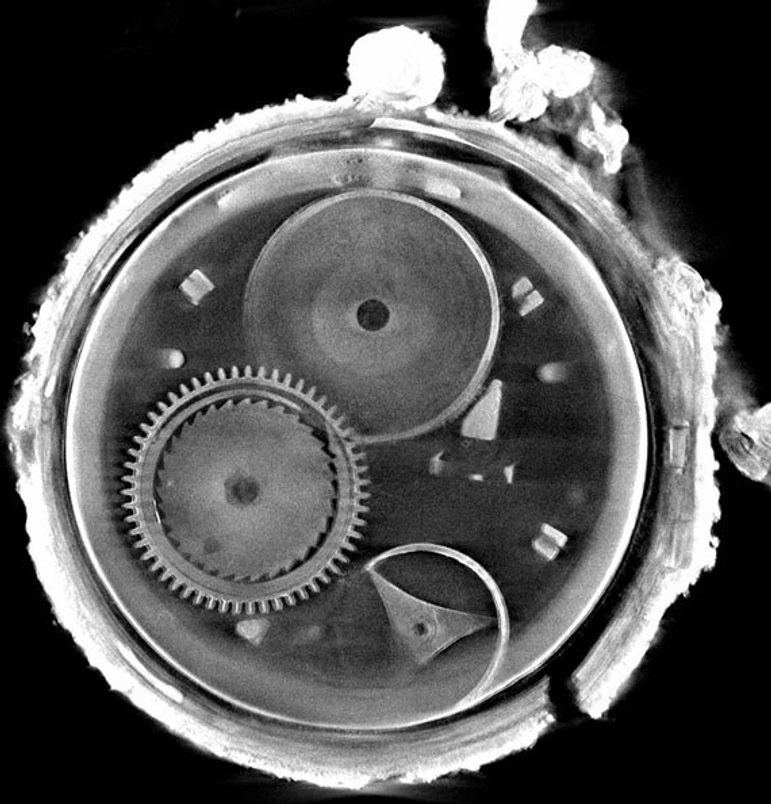 A black and white scan showing the insides of a circular watch from head on, including a cog on the middle left, a circular plate with a hole ion it at the top left, and a round rim with a curved triangular support in the middle. The outside of the watch shows a bumpy outer crust