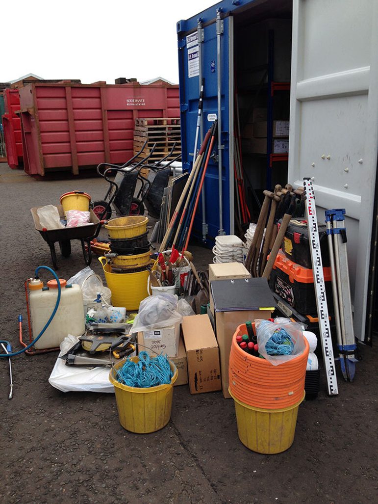 A colour photograph of a lot of digging equipment just outside and open blue outside storage unit area. Equipment including: buckets, shovels, measuring tools, trowels, spray guns, rakes, wheelbarrows and other such items. In the background are red storage units and a pile of wooden pallettes.