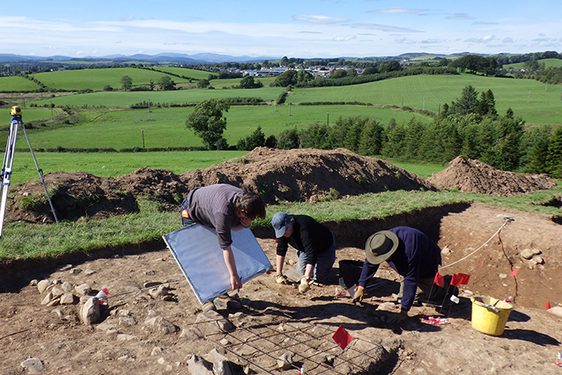 A colour photograph of 3 men in a dug out area in a field. The man to the left is bent over a large wooden board, The other men to the right are on all fours and are digging in the mud. To the far left is a yellow theodolite land measuring instrument on a tall white tripod. Around the edge of the dig are the mounts of earth that had been dug up to create the excavation pit. Behind them are many rolling green fields into the far distance.