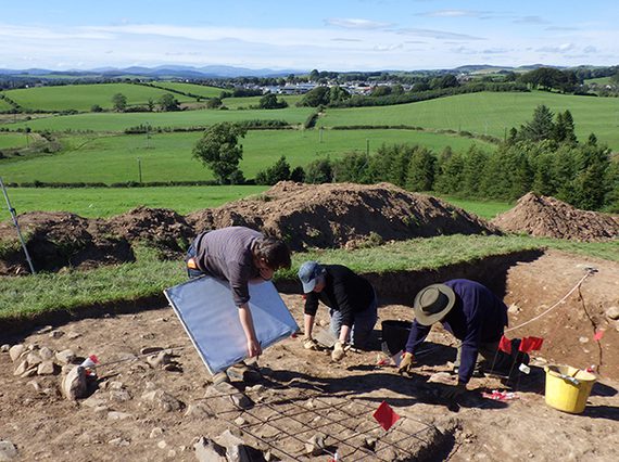 A colour photograph of 3 men in a dug out area in a field. The man to the left is bent over a large wooden board, The other men to the right are on all fours and are digging in the mud. To the far left is a yellow theodolite land measuring instrument on a tall white tripod. Around the edge of the dig are the mounts of earth that had been dug up to create the excavation pit. Behind them are many rolling green fields into the far distance.