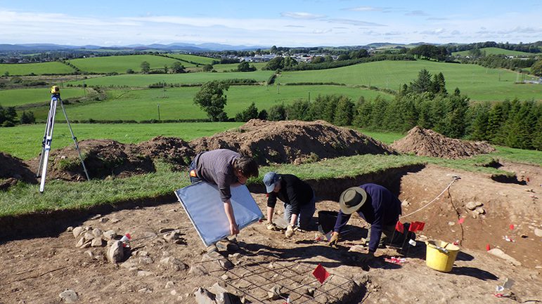 A colour photograph of 3 men in a dug out area in a field. The man to the left is bent over a large wooden board, The other men to the right are on all fours and are digging in the mud. To the far left is a yellow theodolite land measuring instrument on a tall white tripod. Around the edge of the dig are the mounts of earth that had been dug up to create the excavation pit. Behind them are many rolling green fields into the far distance.