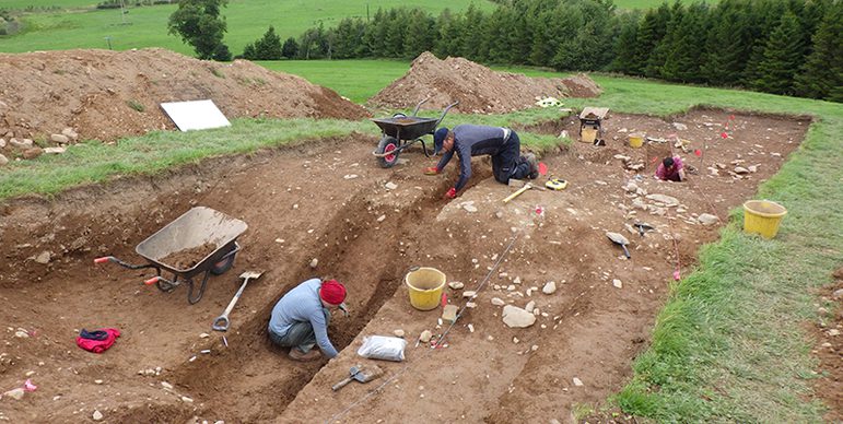 A colour photograph of two people digging in an excavation pit in a large field. In the shallow pit is a much deeper long rectangular sloping ditch. The two people are digging and examining the soil in this pit. To the left are two wheelbarrows full of soil. To the right are a couple of buckets. To the far left are large mounds of soil on the grass of the field. In the background are green fields and trees.