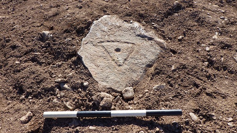 A colour photograph is an oblong rock in the soil with a black and white measuring stick underneath. The rock has a triangle engraved on it with two points at the top and one at the bottom. In the middle of the triangle is a small circular hole dug into the rock.