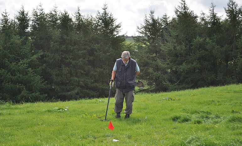 A colour photograph of a man in a very green field with a row of dark green trees behind him. The man is in the centre of shot wearing a short sleeve short and a blue gilet and green army trousers and a cap. He holds a metal detector on his right hand and in front of him is a red flag stuck in the grass.