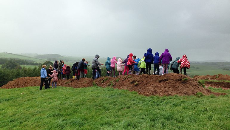 A colour photograph of many schoolchildren on top of an excavation mound of soil in a large green field. They have their backs to us. They are wearing raincoats of many different colours. In the distance, there are hills covered in grey mist.
