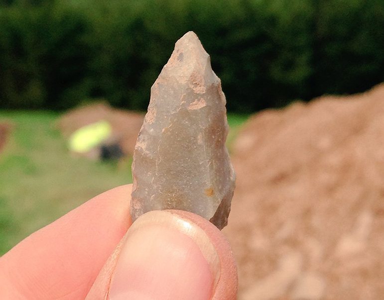A close-up colour photograph of a jagged spear-shaped light brown flint stone held between someone's thumb and forefinger. In the background is a burred pile of soil and a green field and dark green trees.