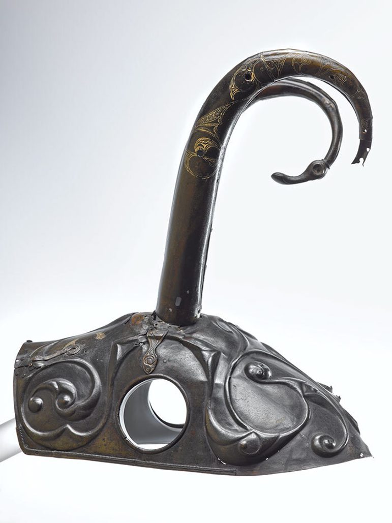 A colour photograph of a bronze horse's headdress with striking curled horns against a white background. The cap part has earholes and is decorated with Celtic swirl patterns. The horns protrude from the centre top and are tall and curved at the ends. One is broken. Te other has a spherical nodule just before the tapered end which looks like an animal head with an eye and a long muzzle.