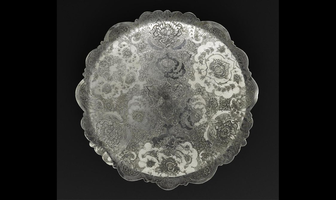 A round silver dish with a scalloped edge with elaborate floral patterns etched on its surface