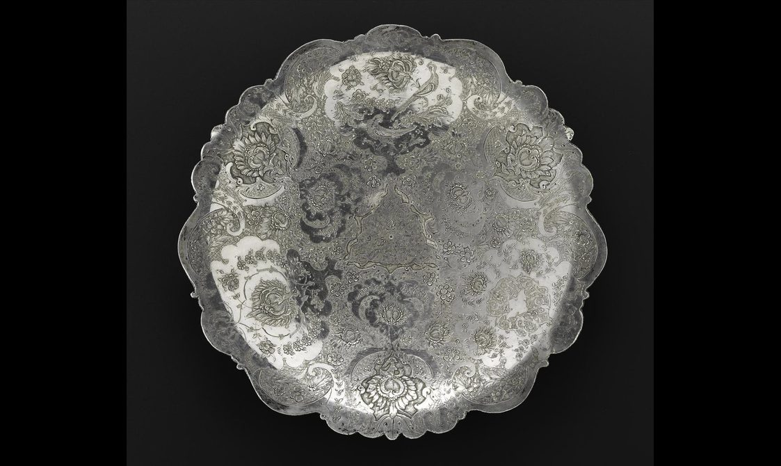 A silver circular dish with a scalloped edge and highly decorated surface.