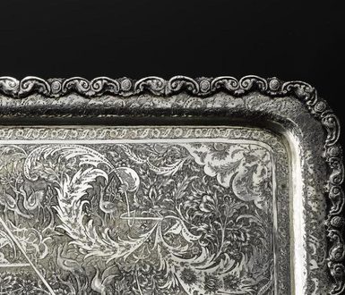 Detail of a silver tray with a decorative border and floral patterns engraved all over its surface.