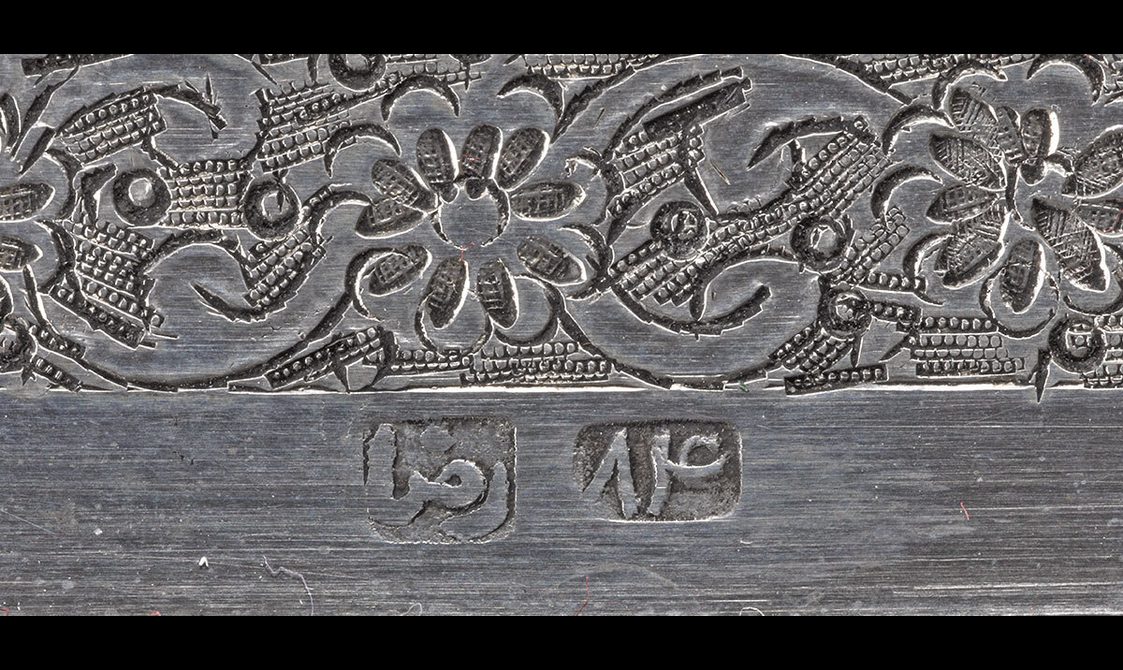 Detail of a silver object with a floral boarder and stamps on its surface to indicate the maker.