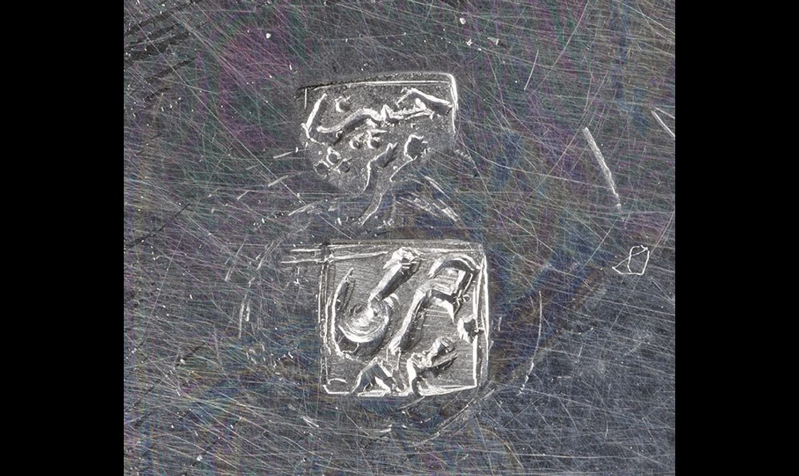 Detail of a silver object with two stamps on its surface indicating the maker.