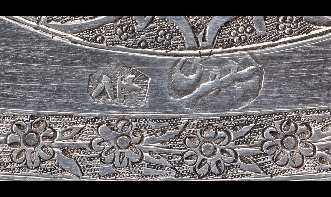 Detail of a silver object with an engraved floral band and two marks noting the maker.