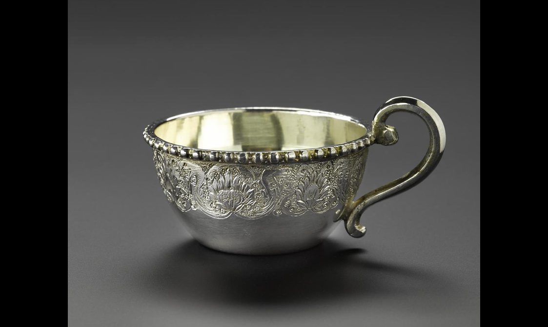 A silver teacup with floral patterns engraved on its surface with a curved handle.