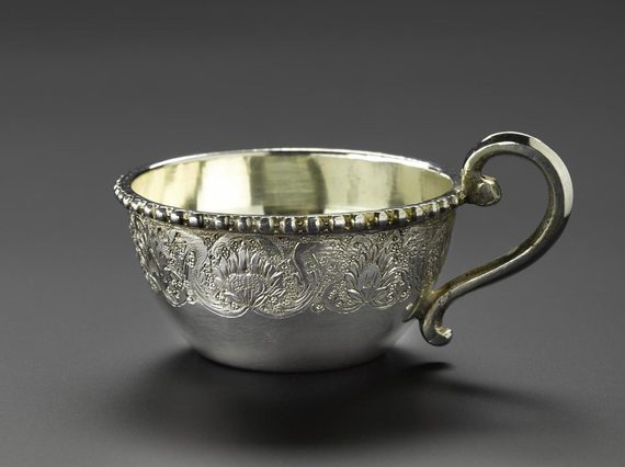 A silver teacup with floral patterns engraved on its surface with a curved handle.