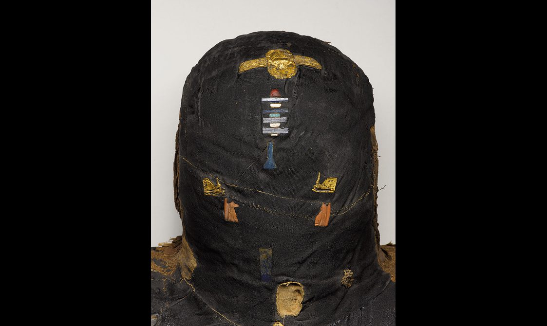Detail of the head of a mummified woman wrapped in blackened bandages with amulets and decorative objects embedded on the surface.