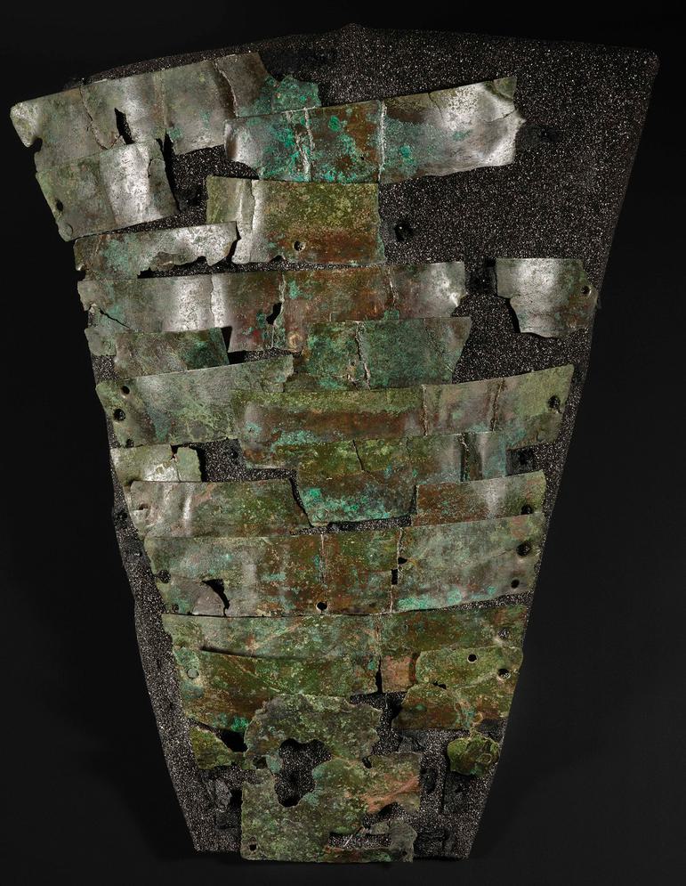 Interlinked series of rusted panel-like metal sheets set upon foam in the rough shape of a limb, getting wider as it goes up.