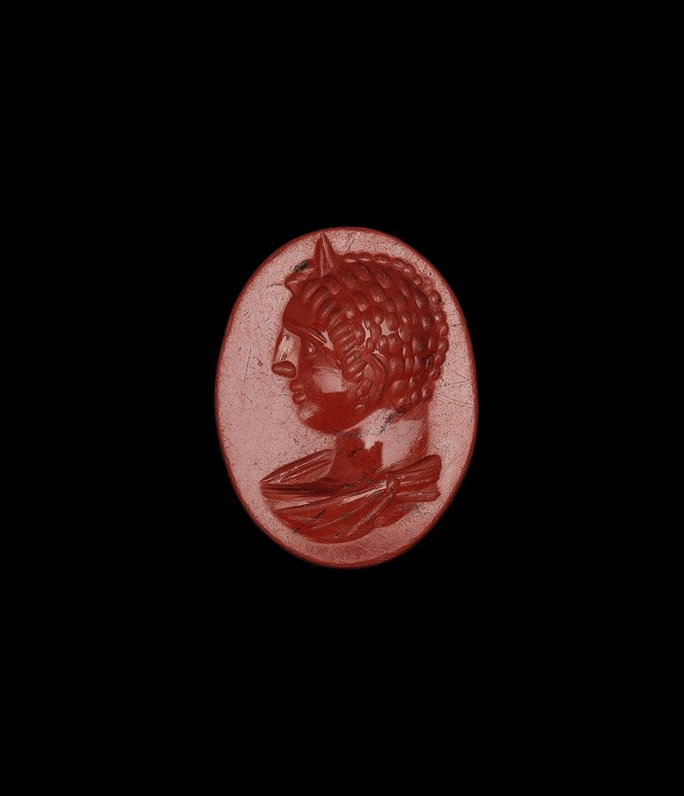 Small red gemstone that looks almost like a wax seal. Egg-shaped, with a male figure sporting right curls and large nose in profile facing left.