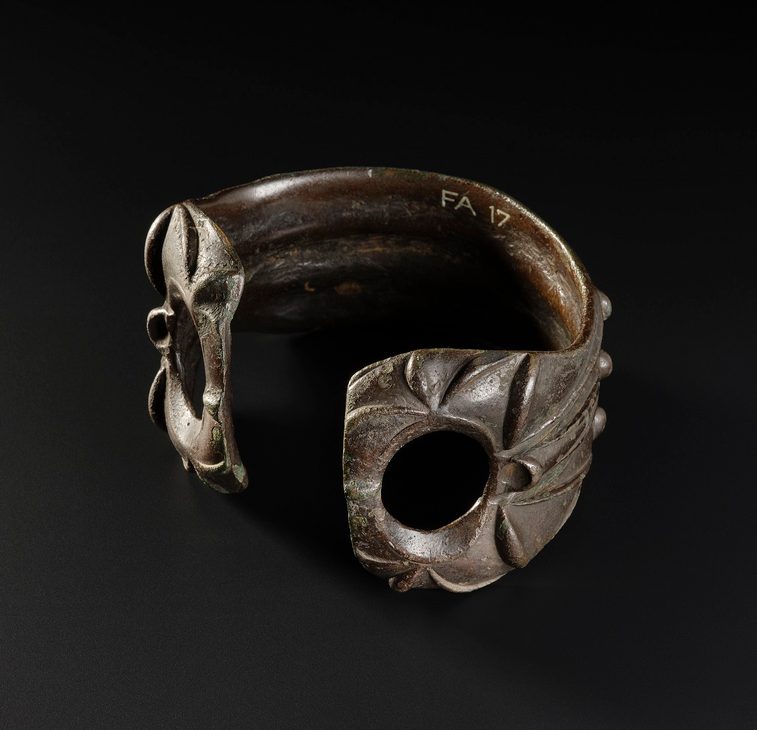 Greenish-brown armlet with an opening facing the viewer. Each end has a large hole in it and a leafy pattern decorates its surface.