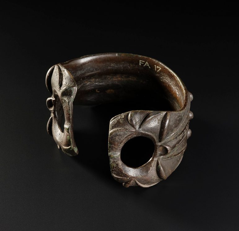 Greenish-brown armlet with an opening facing the viewer. Each end has a large hole in it and a leafy pattern decorates its surface.